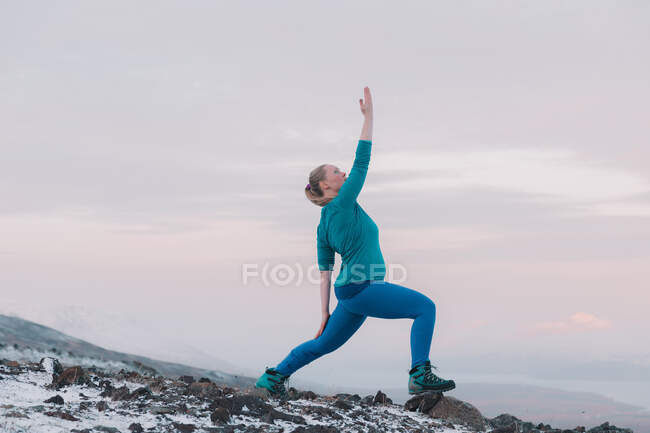 Side view of blond woman with upped leg and hand to side posing on mountain in snow — Stock Photo