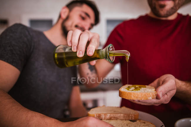 Gay couple pouring olive oil on bread toasts in kitchen — Stock Photo