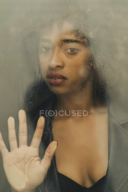 Pretty African American woman looking at camera touching wet glass surface while standing behind window — Stock Photo