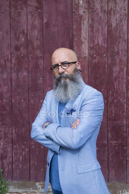 Vista frontal de uma moda madura hipster barbudo em pé contra uma porta vermelha vintage ao ar livre enquanto olha para a câmera — Fotografia de Stock