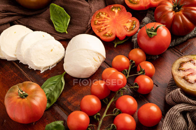 Tomates frescos y queso mozzarella con hojas de albahaca para ensalada en superficie de madera y servilleta de tela - foto de stock