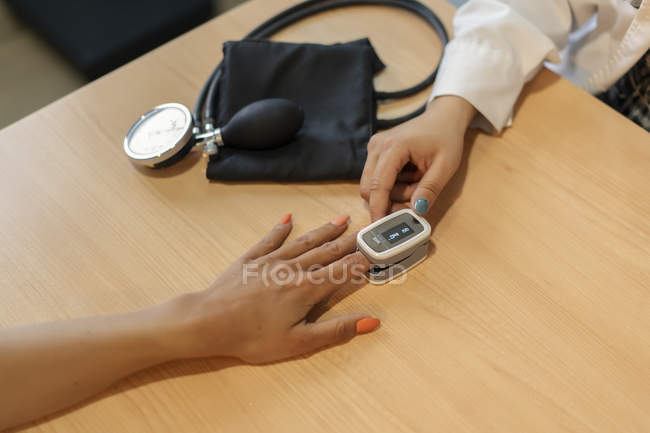 Manos de la doctora y paciente con tonómetro eléctrico en el dedo en la mesa cerca del pulsómetro - foto de stock