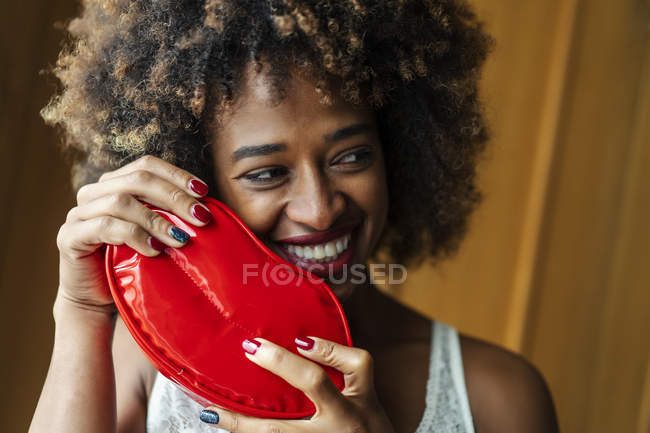 Sonriente hembra negra con el pelo rizado mirando hacia otro lado mientras sostiene la caja en forma de labio brillante en casa - foto de stock