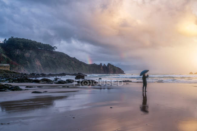 Vista posterior de la persona con paraguas de pie en la orilla del mar alrededor de rocas y salpicaduras de olas al atardecer - foto de stock
