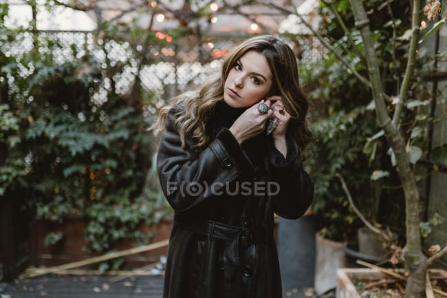 Attractive woman in retro leather coat putting on earrings and looking at camera while standing in small backyard garden — Stock Photo