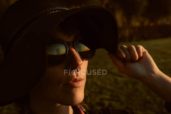 Young sensual brunette wearing black hat and accessories while posing in sunlight with sunglasses — Stock Photo