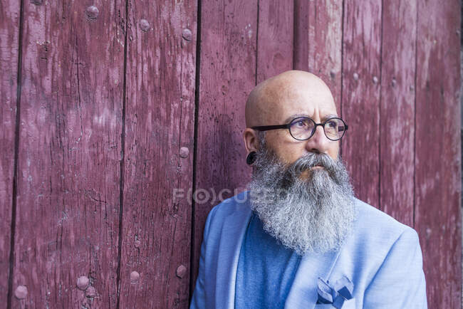 Vista frontal de un hipster barbudo de moda maduro apoyado en una puerta roja vintage al aire libre mientras mira hacia otro lado - foto de stock