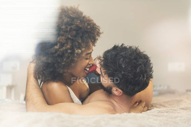 Bearded guy feeding cheerful girlfriend with fresh strawberry while lying on comfortable bed together — Stock Photo