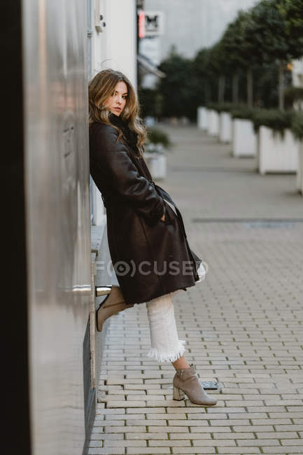 Stylish young woman in vintage leather coat leaning on wall on city street — Stock Photo
