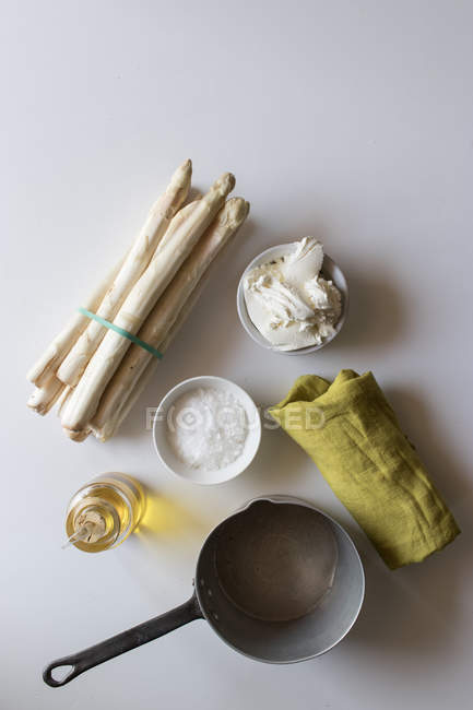 Panela vazia e guardanapo verde na mesa branca perto de um monte de espargos frescos e creme com óleo e sal para cozinhar sopa — Fotografia de Stock