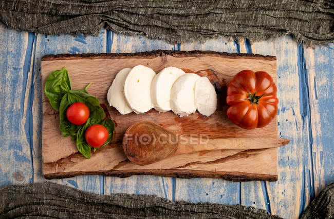 Tomates frescos y queso mozzarella con hojas de albahaca para ensalada caprese rústica sobre tabla de madera - foto de stock
