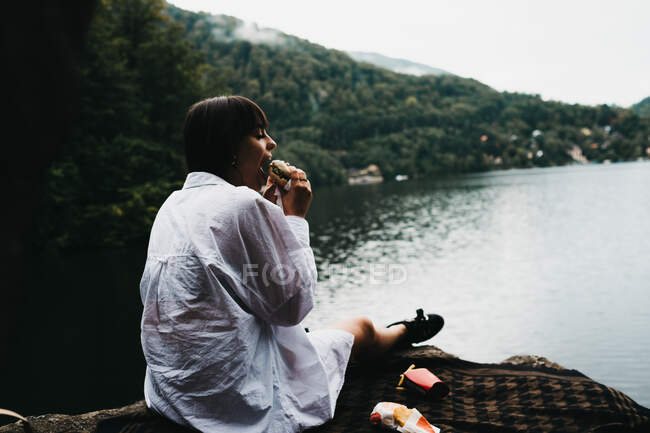 Woman eating hamburger near lake and mountains — Stock Photo