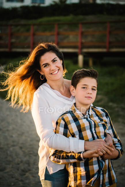 Mujer de mediana edad con su hijo en la orilla del mar sonriendo y abrazándose - foto de stock