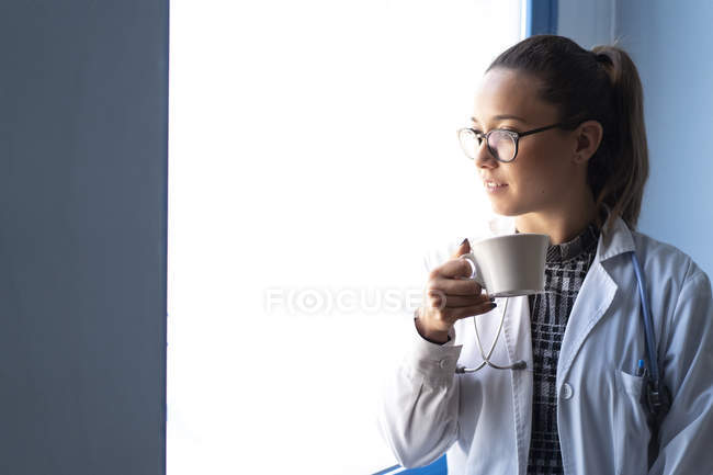 Молодая женщина-врач в форме пьет из чашки в комнате — стоковое фото