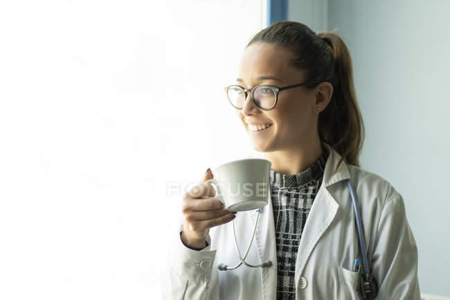 Молода весела жінка-лікар в уніформі пиття з чашки в кімнаті — стокове фото