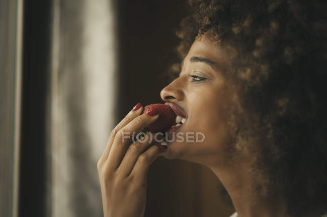 Femme afro-américaine séduisante aux cheveux bouclés mangeant des fraises mûres à la maison — Photo de stock