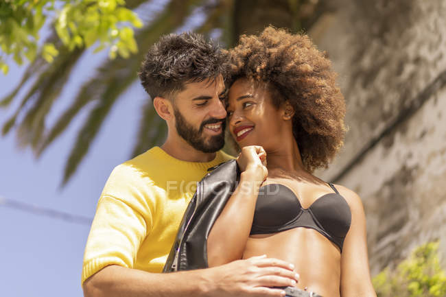 Красивий бородатий хлопець посміхається і фліртує з привабливою чорною жінкою в бюстгальтері, стоячи на міській вулиці разом у сонячний день — стокове фото