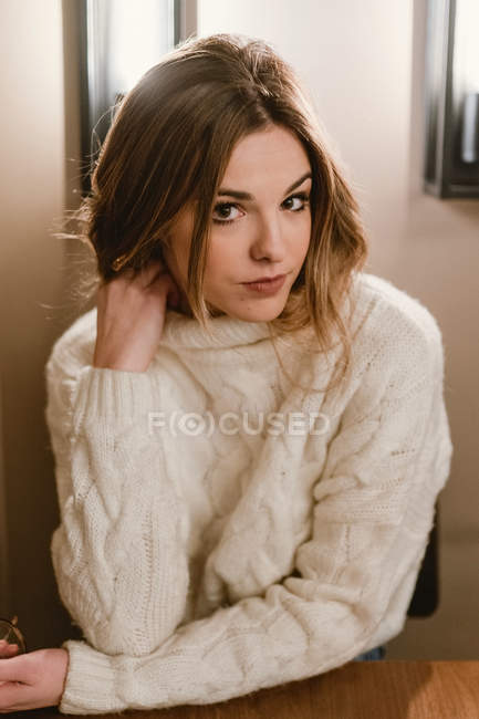 Young female in knitted sweater touching neck and looking at camera while sitting at table in cozy cafe — Stock Photo