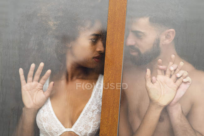 Handsome Hispanic guy touching face to face with seductive African American woman in lace bra while standing behind wet window at home — Stock Photo
