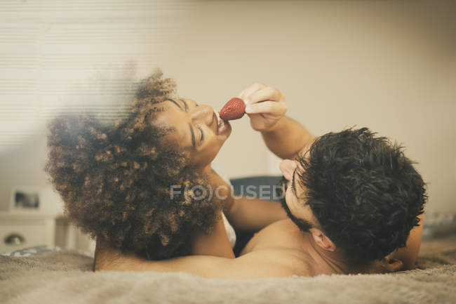 Ragazzo barbuto alimentazione fidanzata allegra con fragola fresca mentre sdraiati su letto comodo insieme — Foto stock
