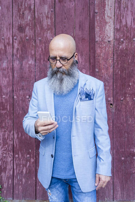 Передній вигляд зрілого модного бородатого хіпстера, що стоїть на червоних старовинних дверях на відкритому повітрі, використовуючи мобільний телефон — стокове фото