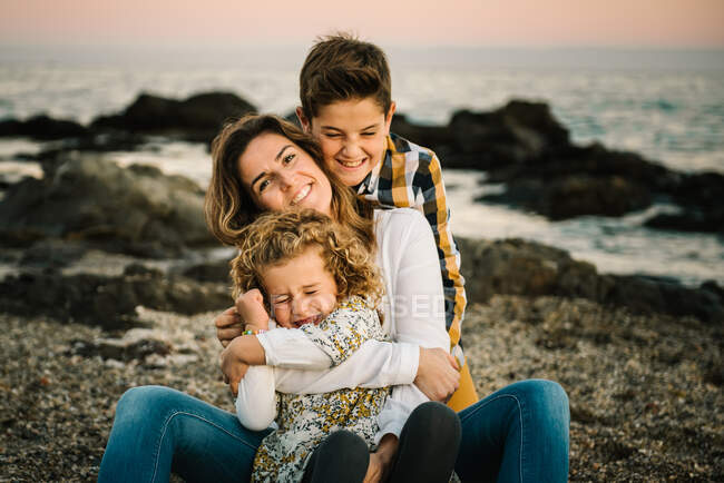 Mujer de mediana edad con sus hijos en la orilla del mar sonriendo y abrazándose - foto de stock