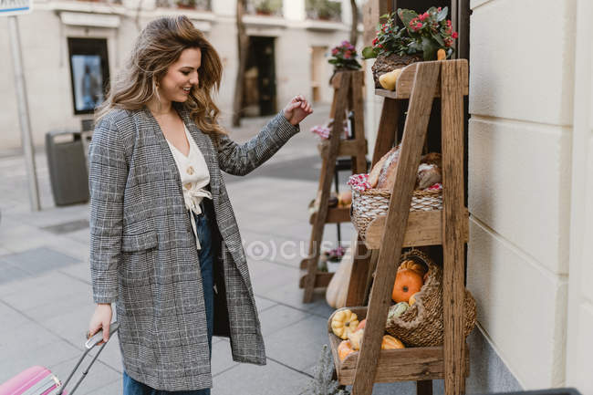 Smiling elegant young woman with suitcase looking at groceries on wooden shelves near store on city street — Stock Photo