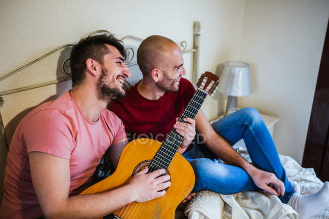 Cheerful gay couple playing guitar in bedroom — Stock Photo