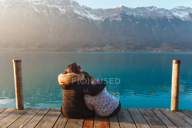 Rückansicht umarmender Frauen auf einem Holzsteg über dem türkisfarbenen See in den schneebedeckten Bergen der Schweiz — Stockfoto