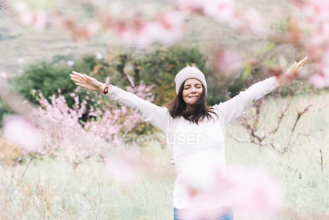 Захоплена жінка в светрі і капелюсі піднімає руки, стоячи в дивовижній весняній сільській місцевості і насолоджуючись свободою — стокове фото
