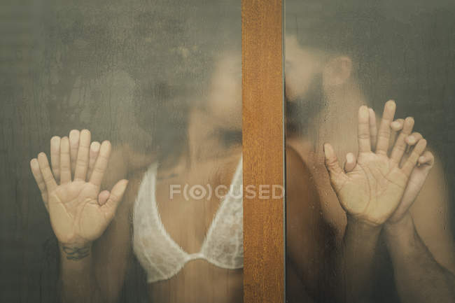 Handsome Hispanic guy touching and kissing seductive African American woman in lace bra while standing behind wet window at home — Stock Photo