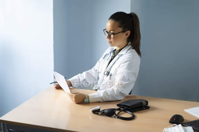 Junge Ärztin in Uniform schaut auf Papier und sitzt am Tisch mit Tonometer im Zimmer — Stockfoto