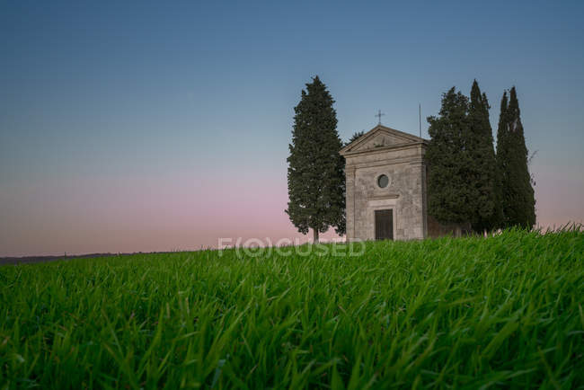 Тихий ландшафт малої каплиці з кипарисами у віддаленому порожньому зеленому полі на заході сонця в Тоскані, Італія — стокове фото