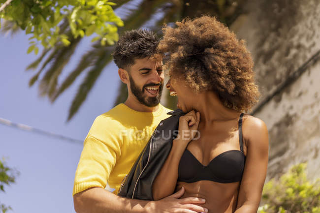 Красивый бородатый парень улыбается и флиртует с привлекательной черной женщиной в лифчике, стоя вместе на городской улице в солнечный день — стоковое фото