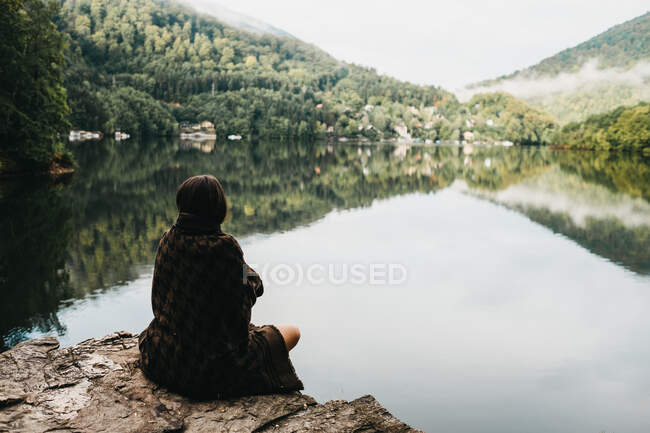 Женщина сидит с одеялом возле озера и гор — стоковое фото