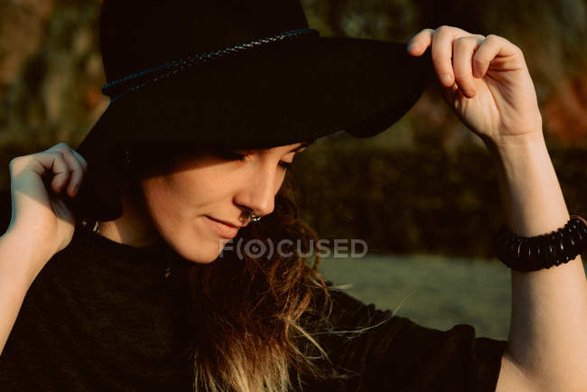 Jovem morena sensual vestindo chapéu preto e acessórios enquanto posando na luz solar mantendo os olhos fechados — Fotografia de Stock