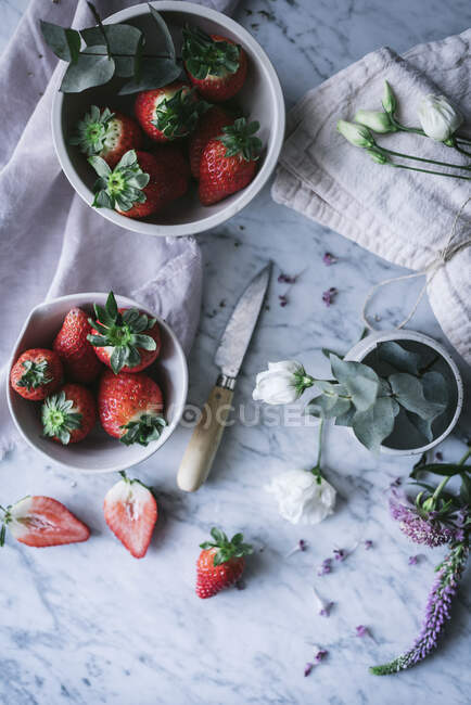 Bowls with ripe strawberries and beautiful flowers placed on white marble tabletop near knife — Stock Photo