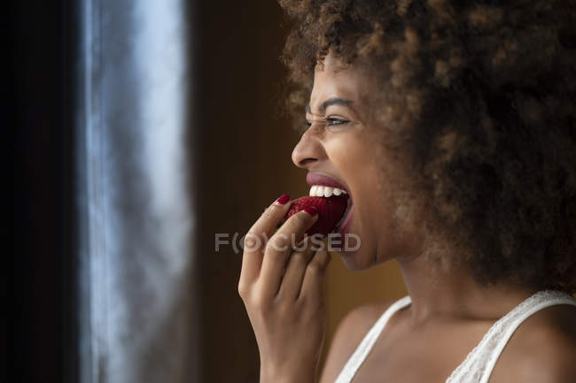 Mulher afro-americana sedutora com cabelo encaracolado comendo morango maduro em casa — Fotografia de Stock