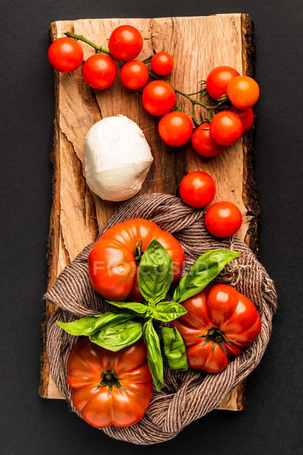 Frische reife Tomaten, Mozzarella und Basilikumblätter auf einem Stück Holz auf schwarzem Hintergrund — Stockfoto