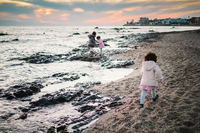 Anonymous little girls from behind standing by the beach on rocks looking at the sea on a winter day — Stock Photo