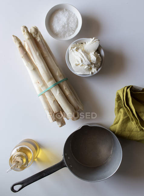 Cazo vacío y servilleta verde sobre mesa blanca cerca de manojos de espárragos frescos y crema con aceite y sal para cocinar sopa - foto de stock