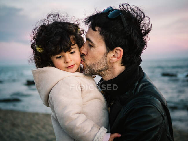 Niedliche Szene, wie Papa ihre kleine Tochter im Winter am Strand hält und küsst — Stockfoto