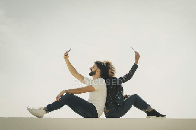 Young multiethnic man and woman smiling and taking selfies while sitting back to back on building wall against gray sky — Stock Photo