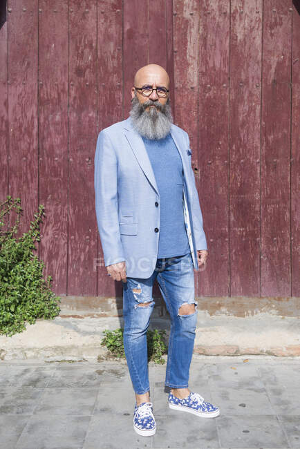 Vista frontal de un hipster barbudo de moda maduro de pie contra una puerta roja vintage al aire libre mientras mira a la cámara - foto de stock