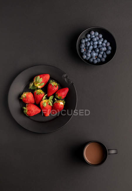 Várias bagas frescas e caneca de bebida quente aromática para o café da manhã no fundo preto — Fotografia de Stock