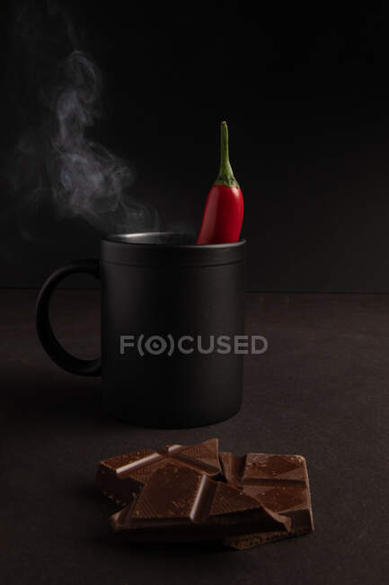 Leckere Schokoladenstücke in der Nähe eines schwarzen Bechers dampfenden Heißgetränks mit Chilipfeffer auf dunklem Hintergrund — Stockfoto