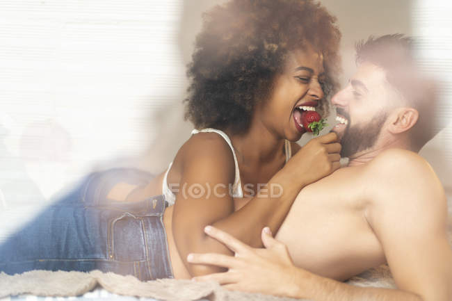 Бородатый парень кормит веселую подружку свежей клубникой, лежа на удобной кровати вместе — стоковое фото