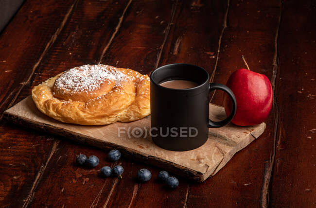 Frischer roter Apfel und reife Blaubeeren auf Holztisch neben Becher mit aromatischem Heißgetränk und leckerem Brötchen zum Frühstück am Morgen — Stockfoto