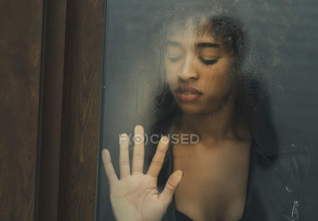 Jolie femme afro-américaine avec les yeux fermés touchant surface en verre humide tout en se tenant derrière la fenêtre — Photo de stock