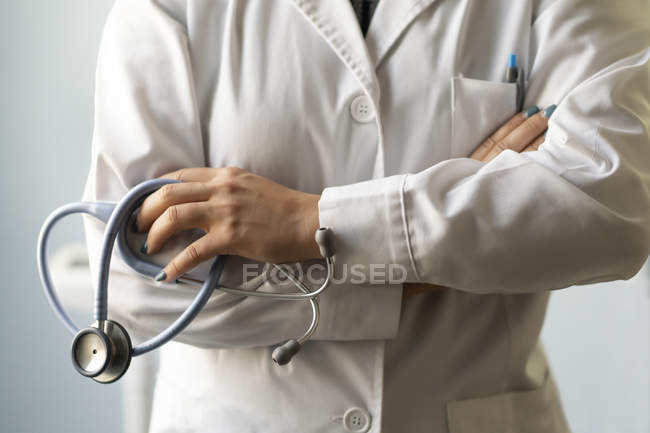 Primer plano de la doctora en uniforme con las manos cruzadas sosteniendo estetoscopio en la habitación - foto de stock